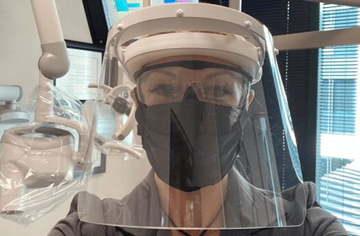 The image shows a photo of a woman wearing a Face Shield in a dental lab as apart of her PPE.